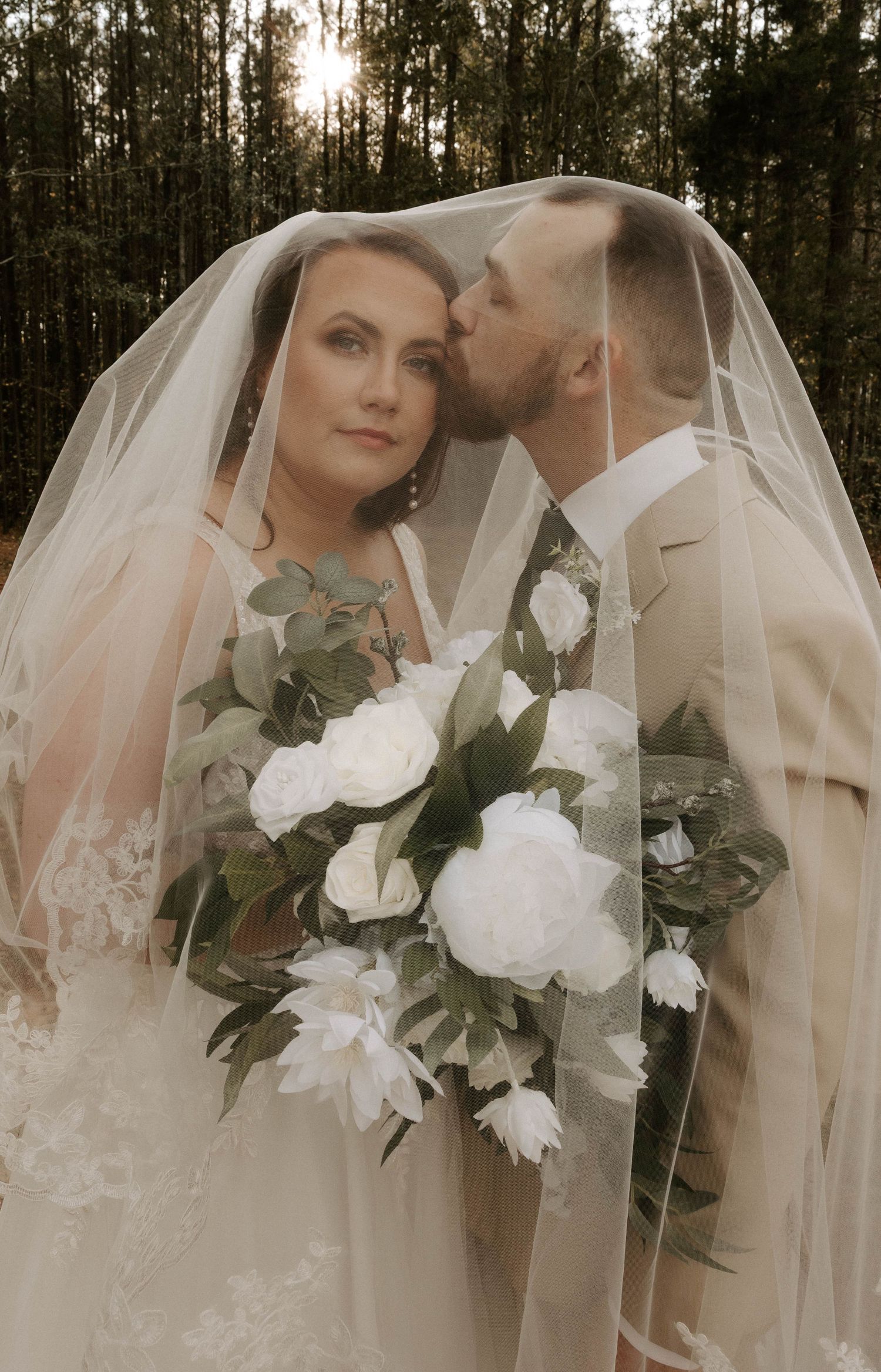 A romantic wedding portrait under a veil with a beautiful white floral bouquet in a forest setting at sunset.