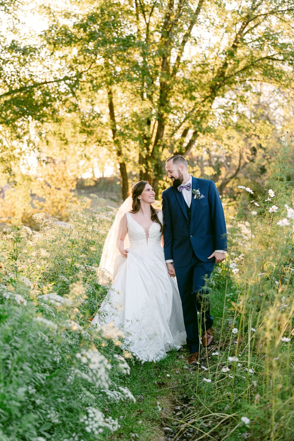 A couple in wedding attire walks together through a dreamy meadow filled with wildflowers at golden hour.