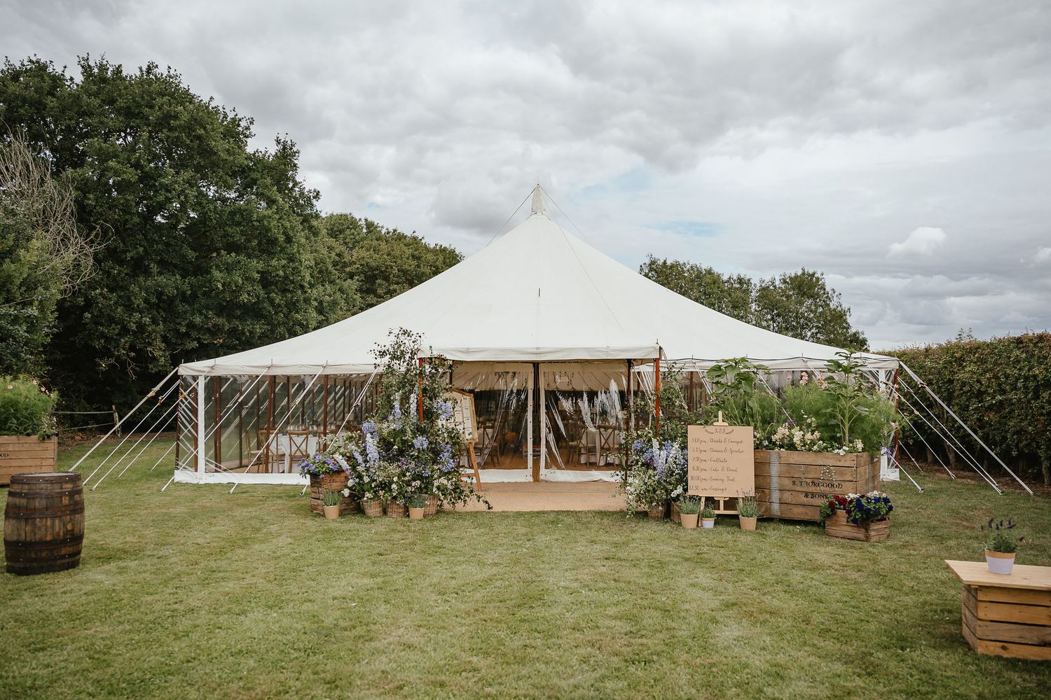 A white wedding tent set up on a green lawn with wooden barrels and floral decorations at the entrance.