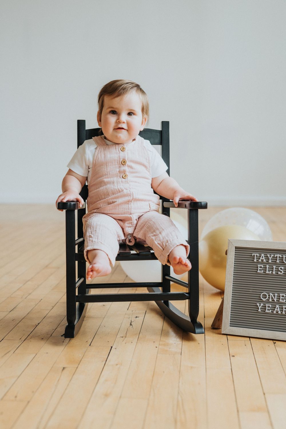 A small child sits in a black rocking chair on hardwood floors next to a letterboard in a minimalist setting.