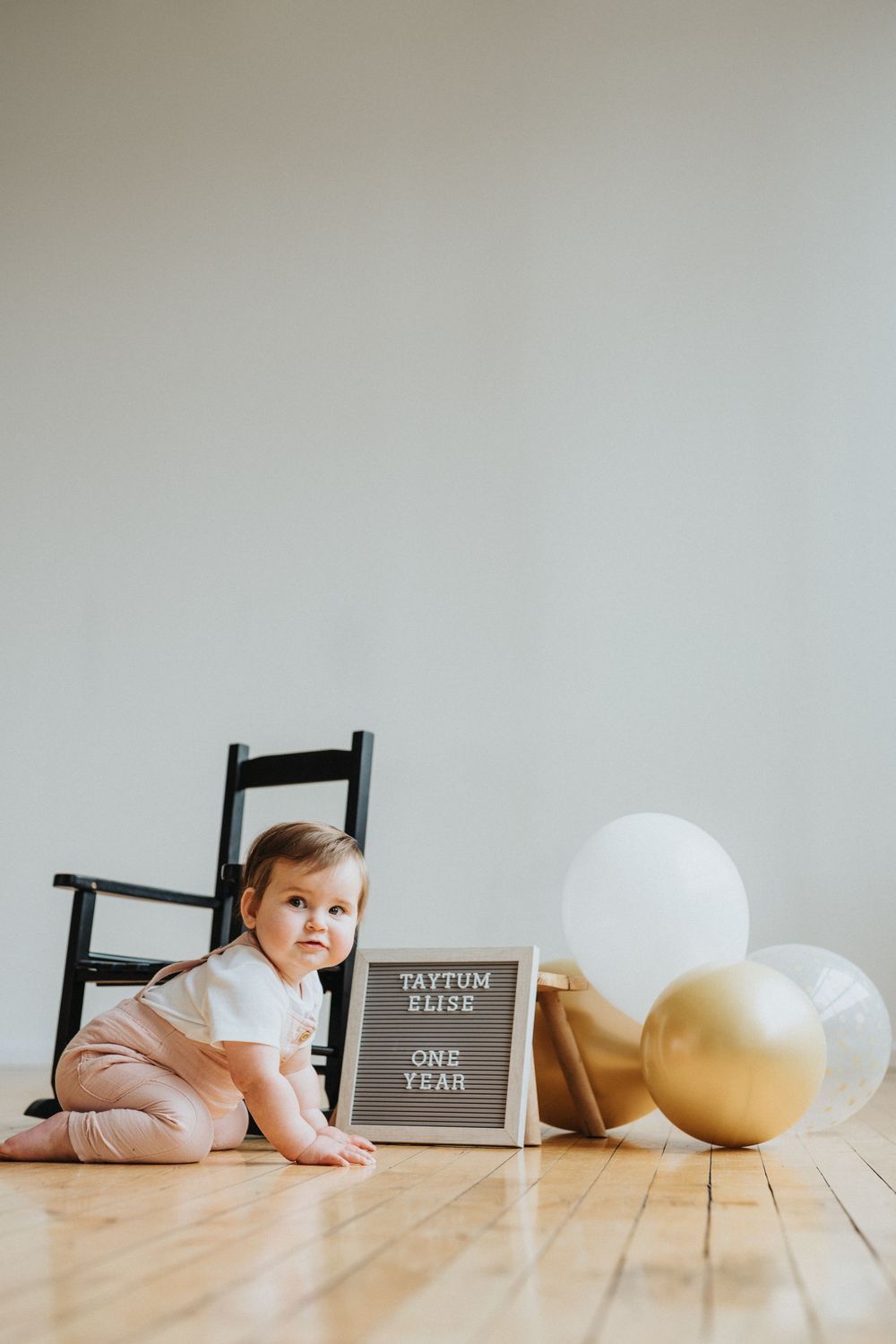 A baby in pink clothing lies on a wooden floor near a letter board and gold balloon in a minimalist setting.