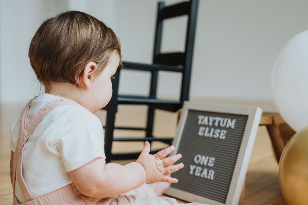 Baby in white outfit sitting next to black and white milestone cards on wooden floor.