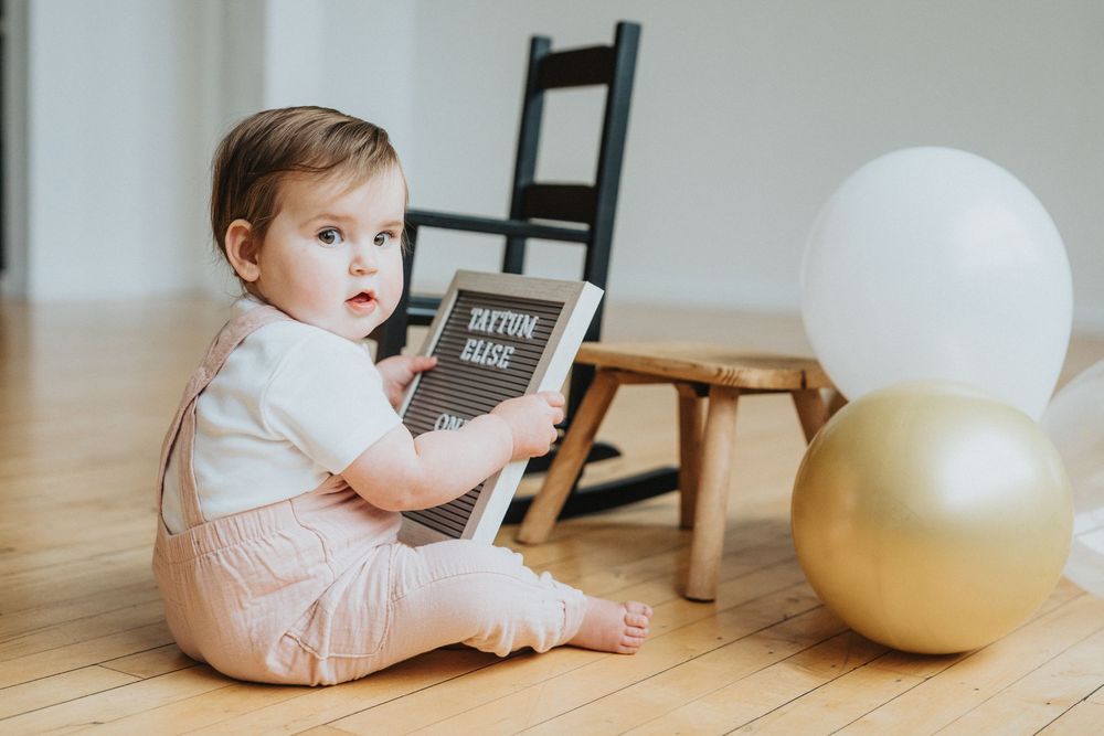 Baby in white outfit sits on wooden floor playing with large exercise ball in bright room.