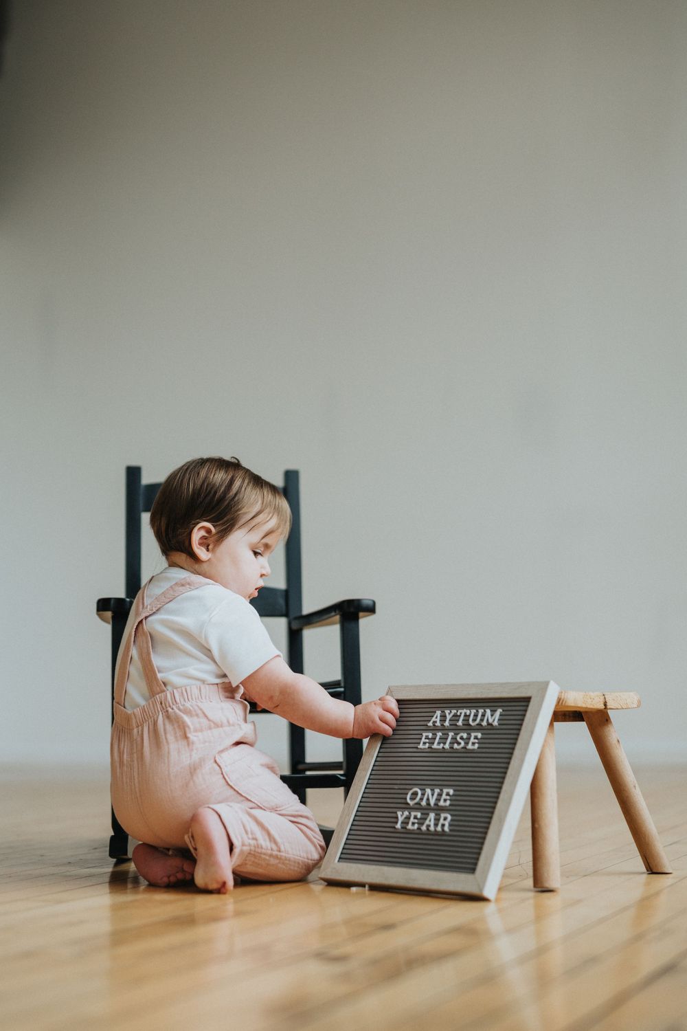 Baby in white onesie crawls on wooden floor examining milestone chalkboard signs showing growth progress.