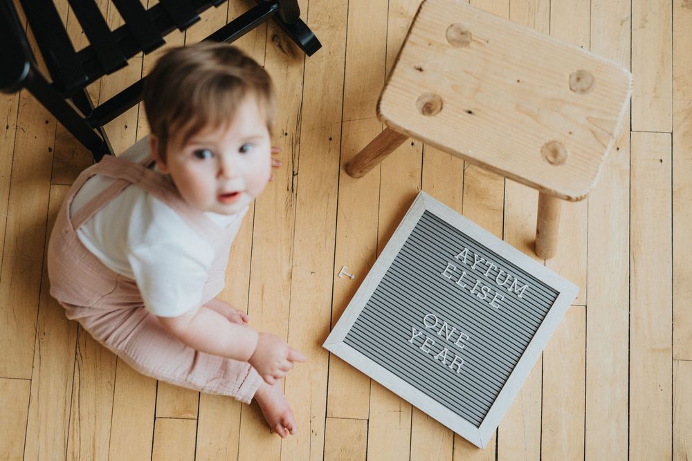 Baby on wooden floor reaching towards gray letter board displaying milestone message.