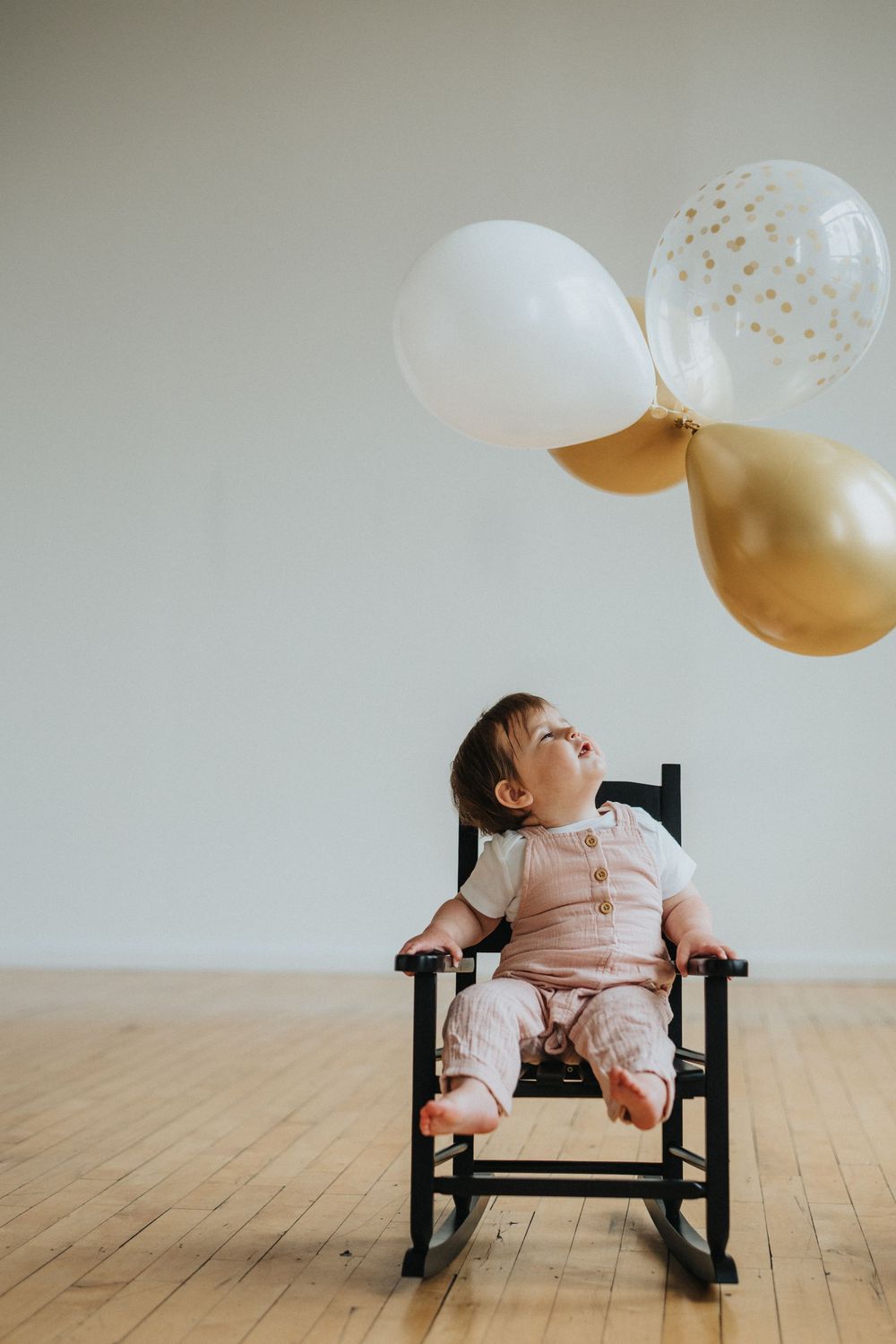 A baby in pink pajamas sits in a black rocking chair with white and gold balloons floating overhead in a minimalist setting.