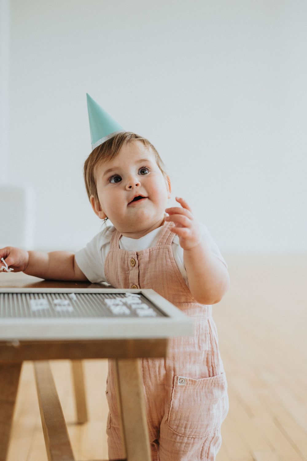 A baby in a striped romper crawls and plays around a wooden table on a hardwood floor.
