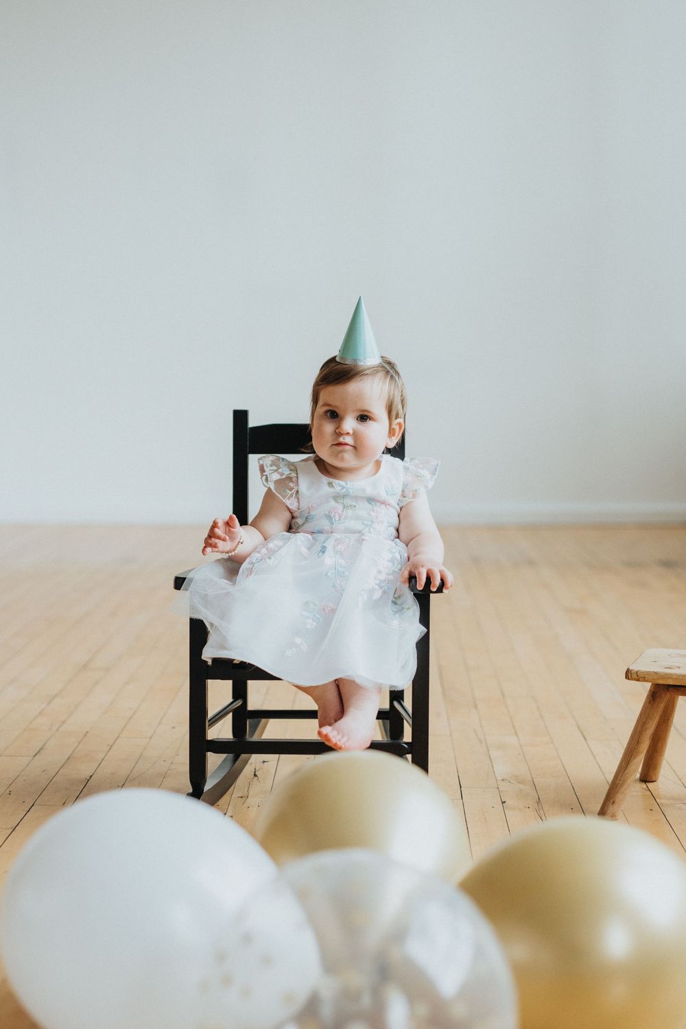 Baby in white dress sits in black chair with party hat and white balloons for first birthday celebration.