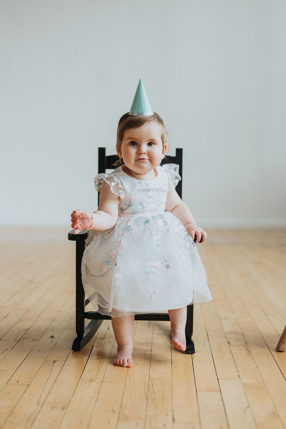 A small child in a white floral dress sits in a dark wooden chair on a hardwood floor.
