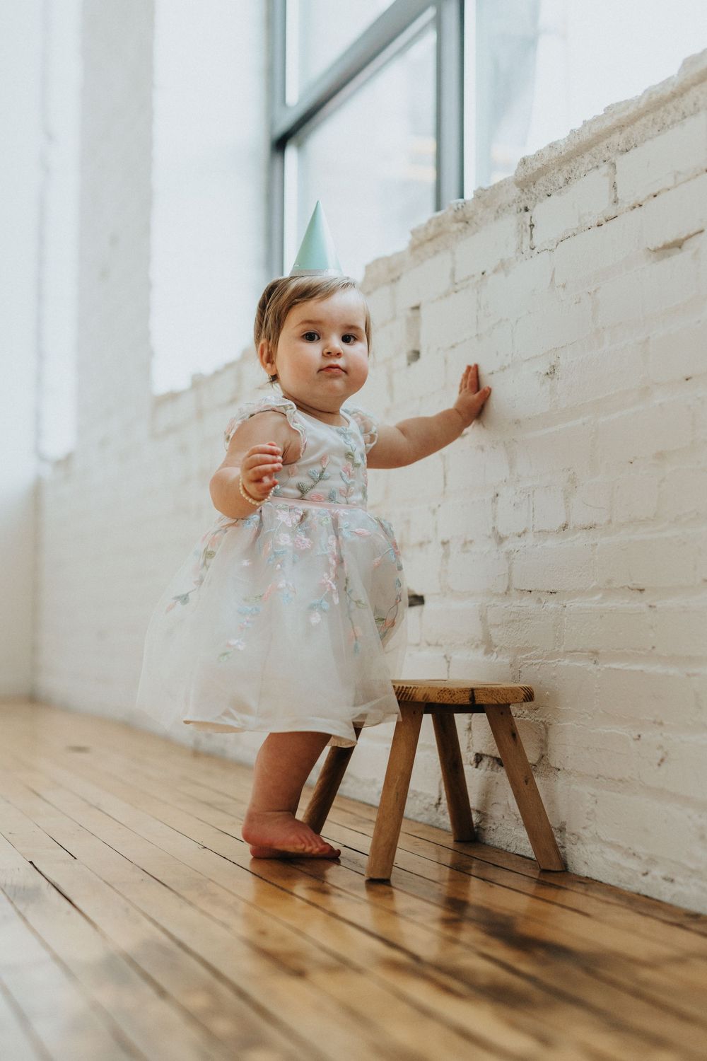 Baby in white dress explores wooden floor near rustic white brick wall during birthday photoshoot.