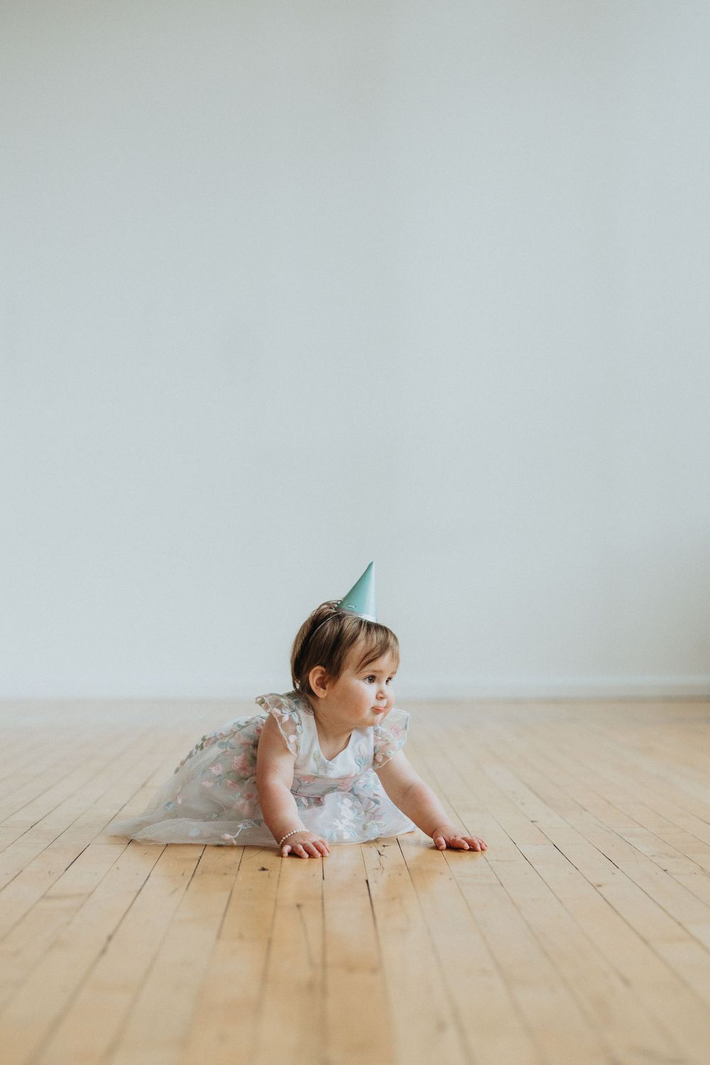 Young child in white dress crawls across hardwood floor in minimalist setting with natural light.