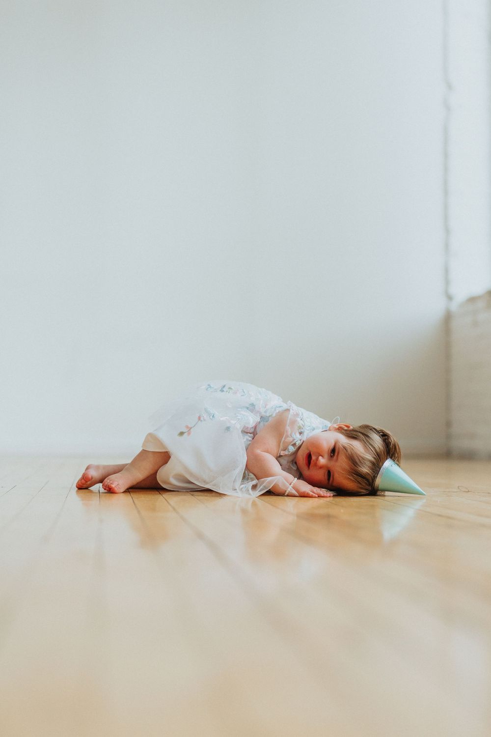 A small figure in white clothing lies curled up on a hardwood floor beside a mint green pillow.