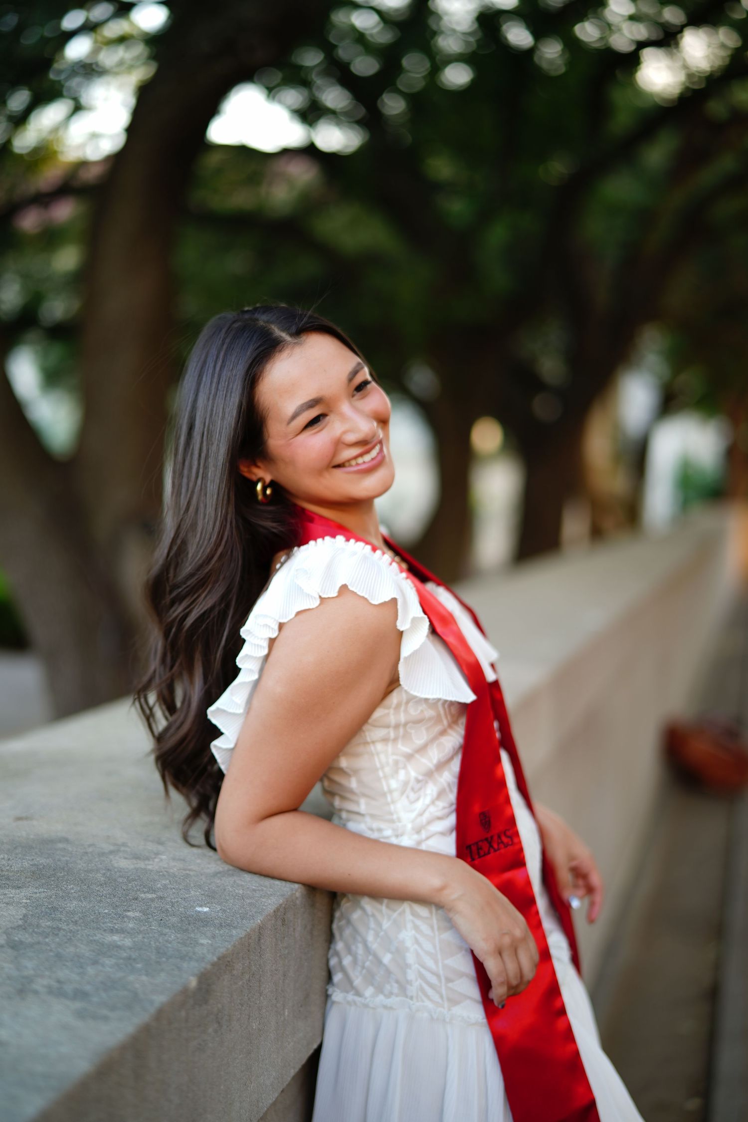 A person in a white lace dress and red sash smiles while posing outdoors in a natural setting.