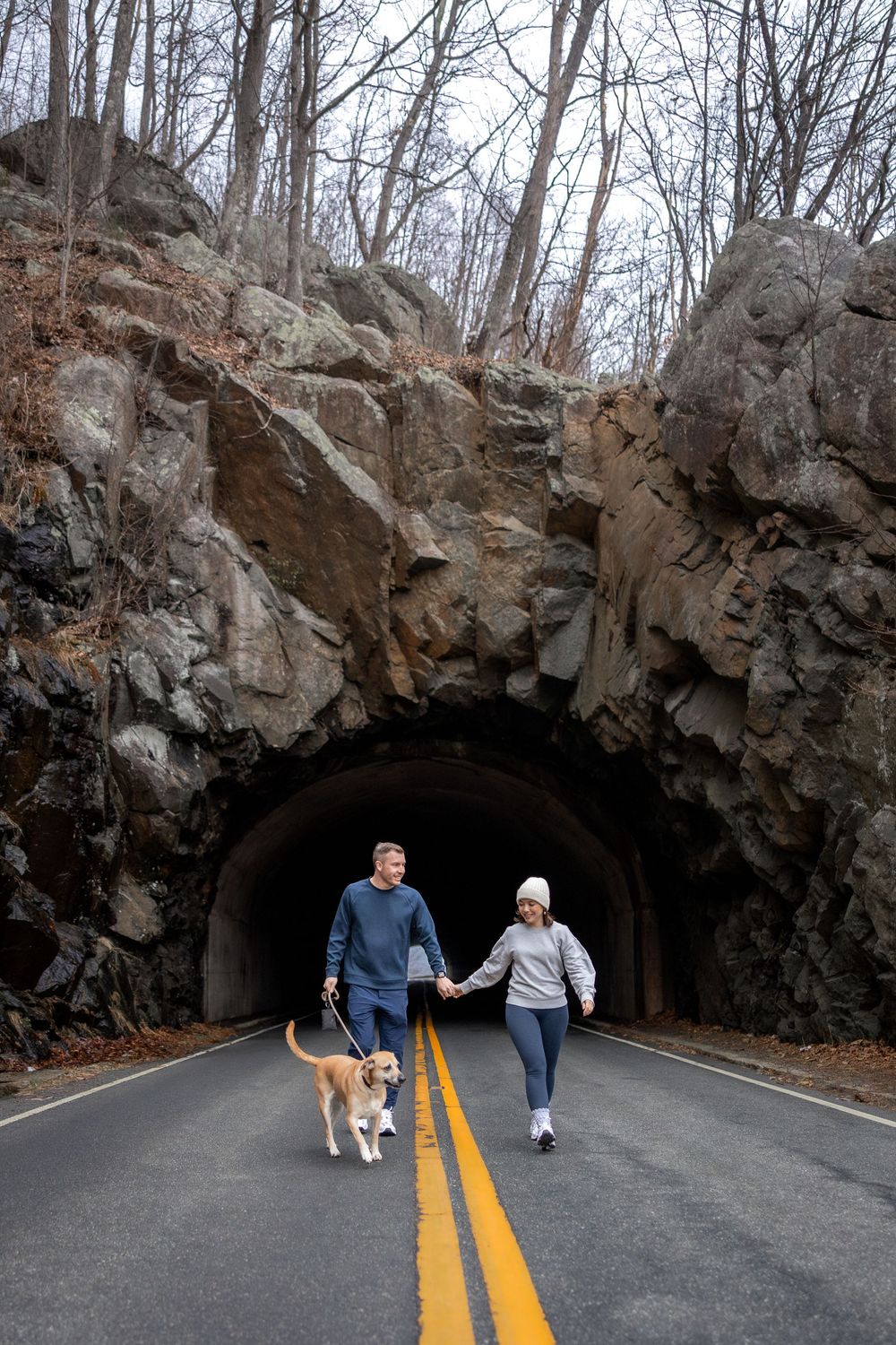 A couple walks their dog through a scenic rock tunnel carved into a mountainside on a paved road with yellow lines.