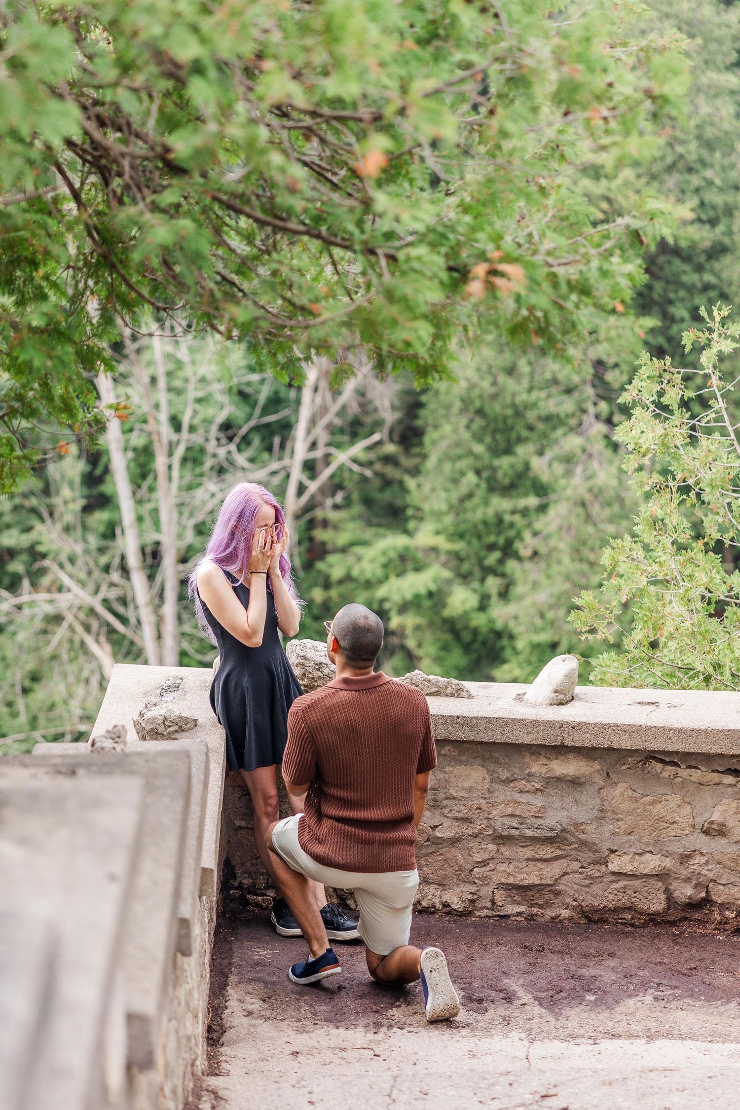 Two people sit together on a stone wall overlooking a scenic wooded area on a bright summer day.