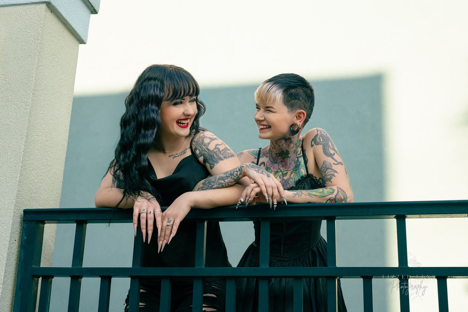 Two tattooed women in black dresses smiling on a balcony.
