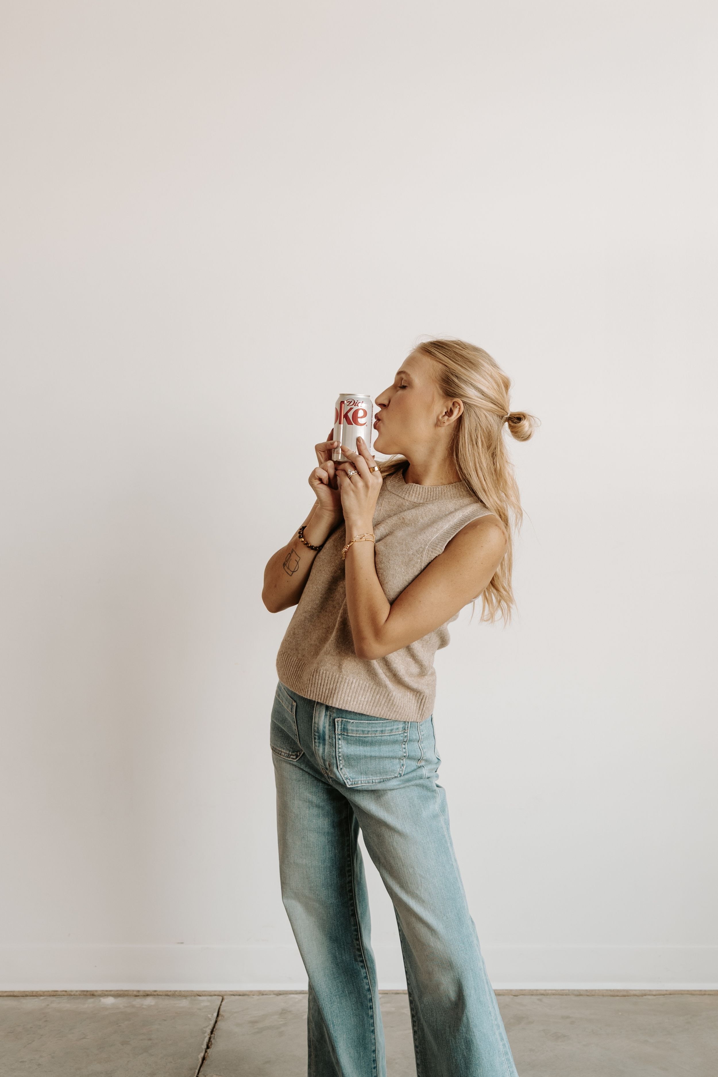 Woman in beige sweater and jeans dancing with a coffee cup against white wall.