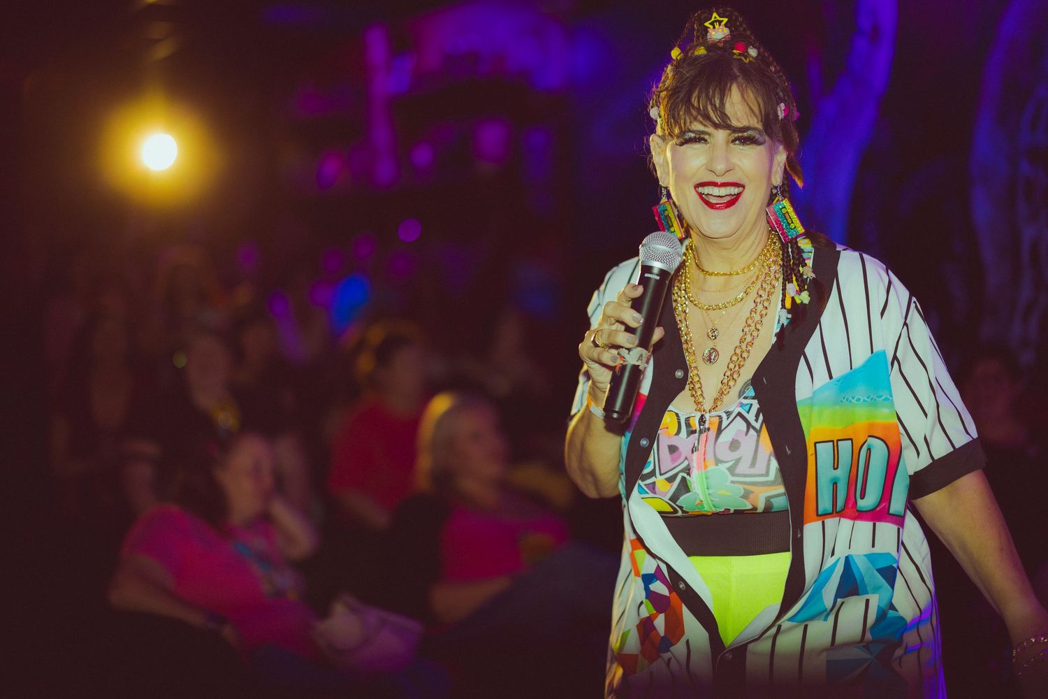 Energetic performer in colorful retro outfit holds microphone on stage before a lively crowd at a vibrant nighttime event.