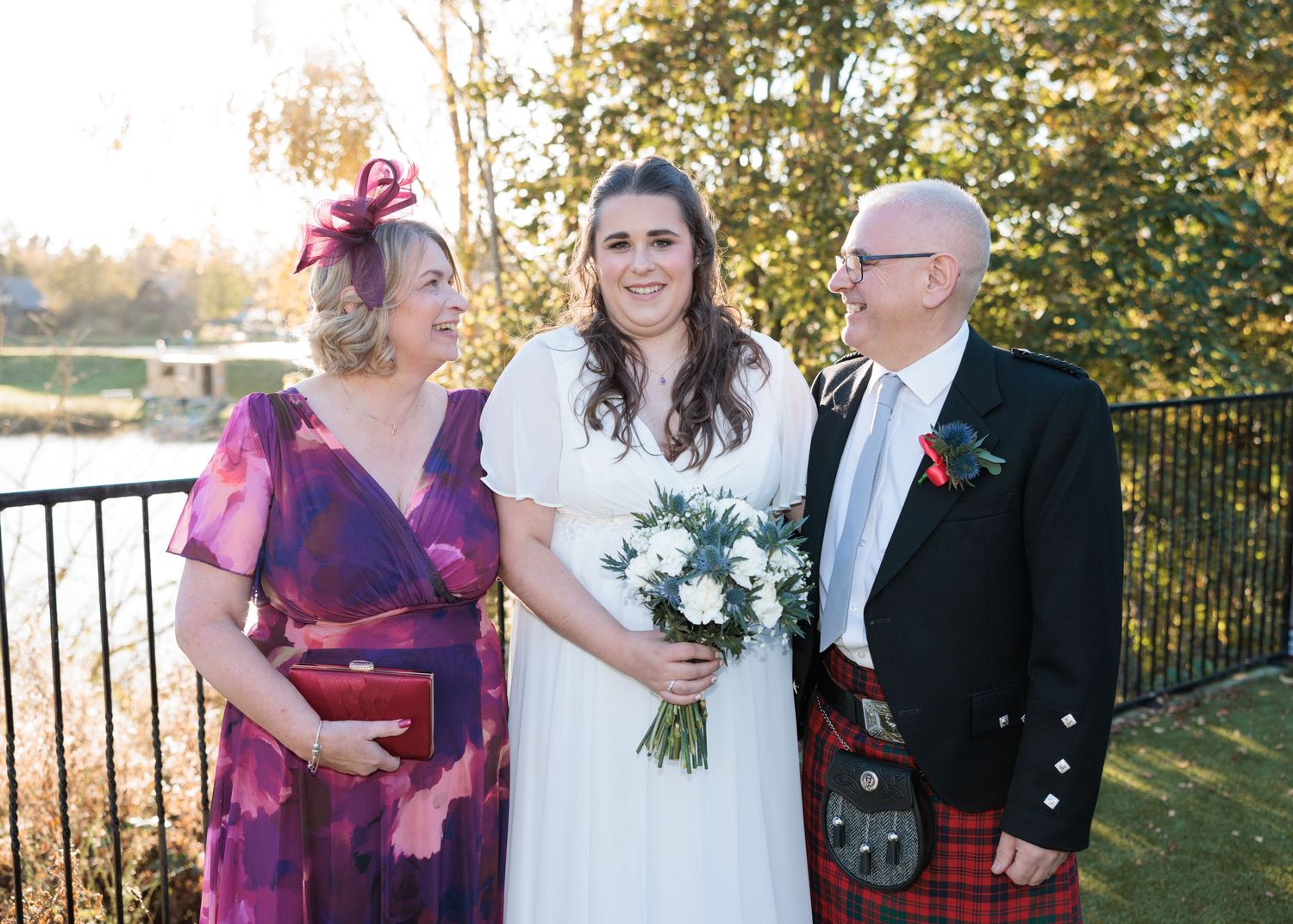 Wedding party in burgundy and formal attire poses outdoors on a sunny autumn day with trees in the background.