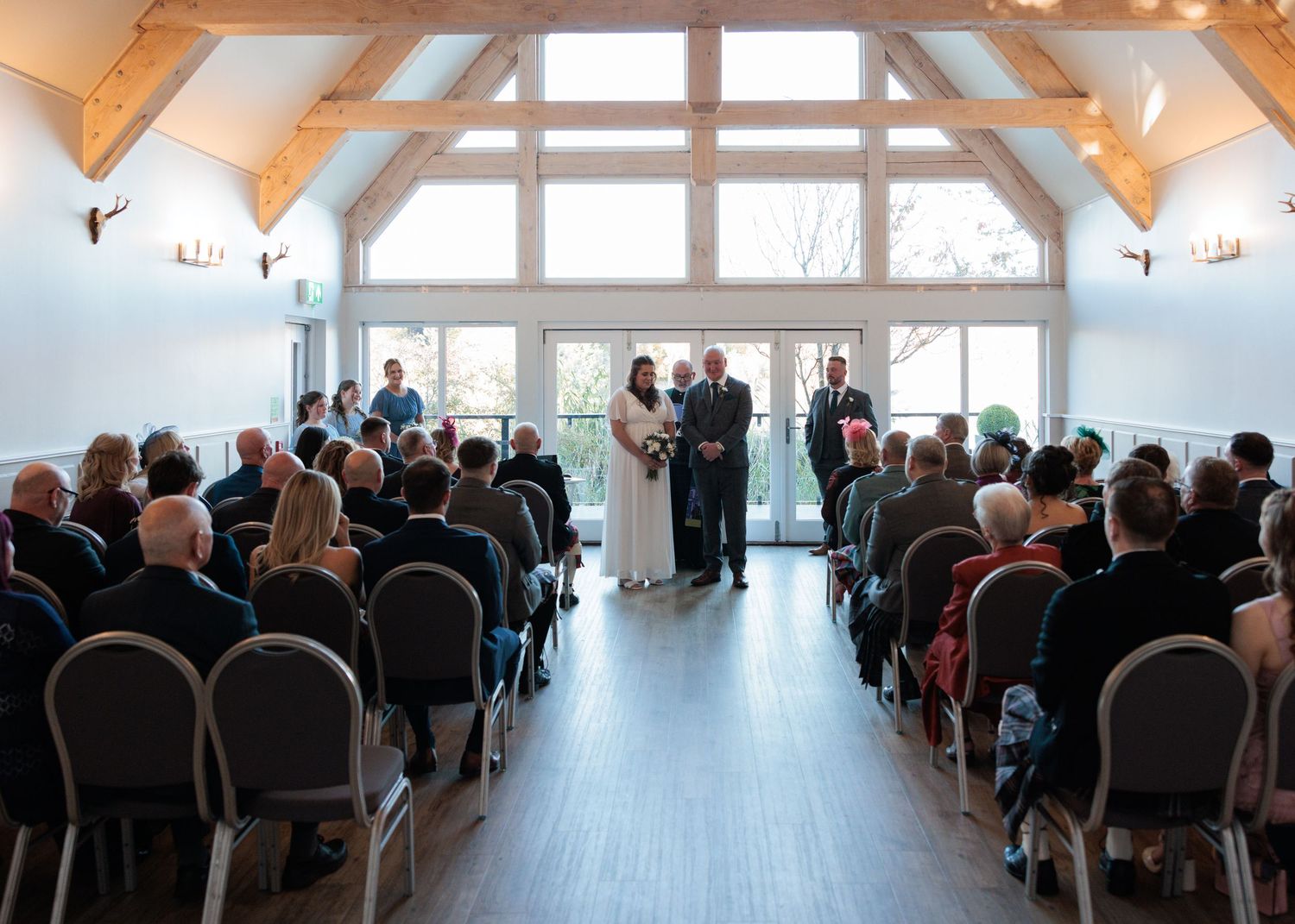 Indoor wedding ceremony in a bright venue with vaulted wood beam ceiling and large windows overlooking a scenic view.