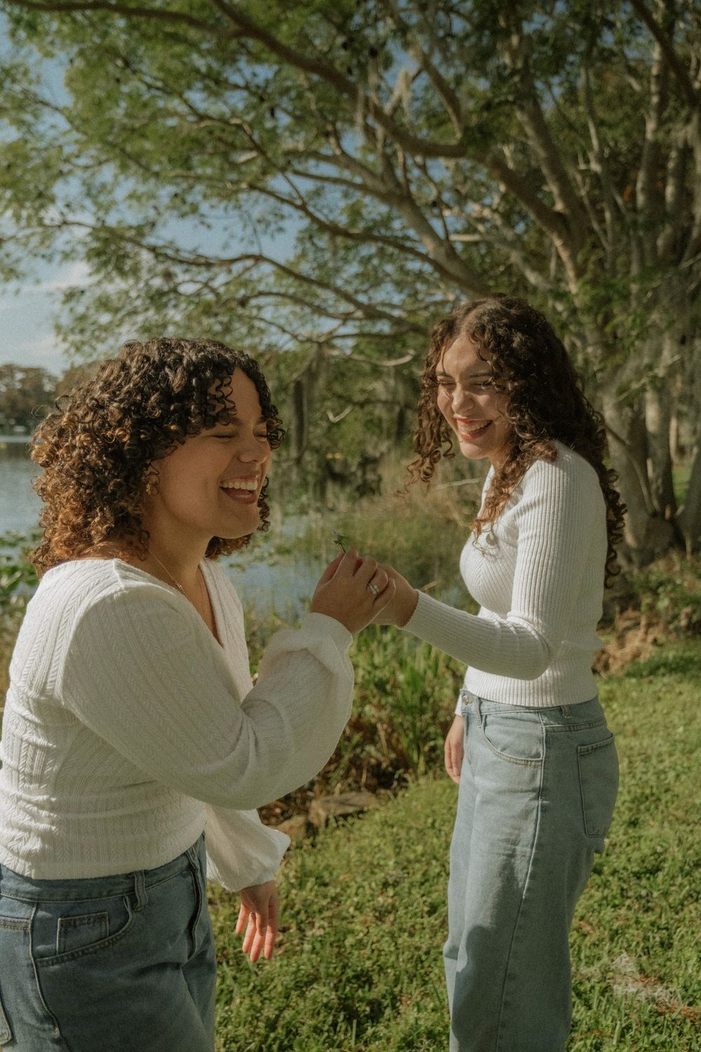 Two friends sharing a joyful moment outdoors with trees and water in the background.