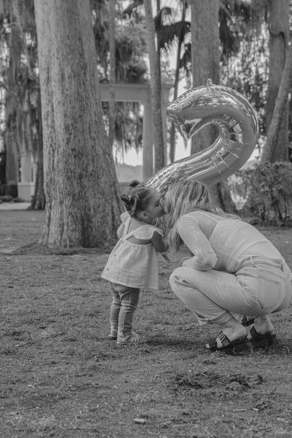 A black and white photo showing a tender moment between a child and adult with a number 2 balloon in a wooded setting.