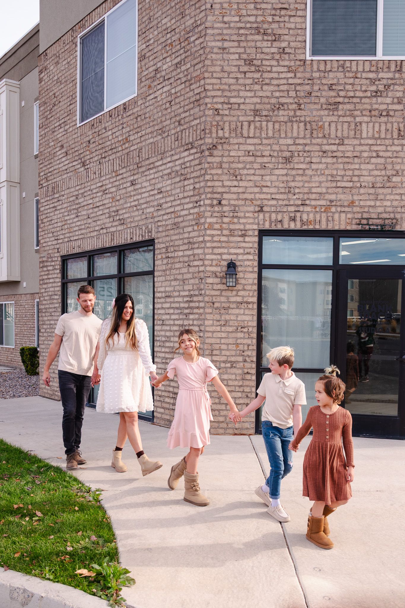 Family of five walking hand in hand outside modern brick building on sunny day.