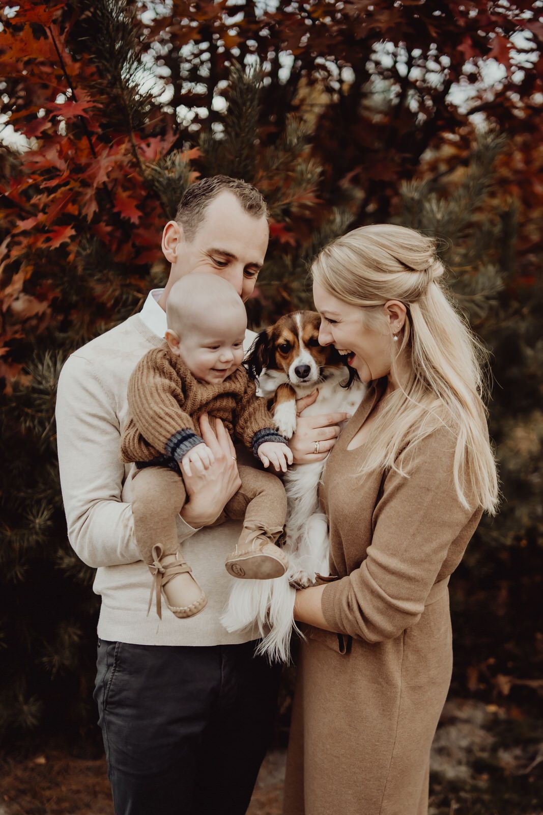 A couple shares a tender moment outdoors with their baby and small dog against a backdrop of autumn foliage.