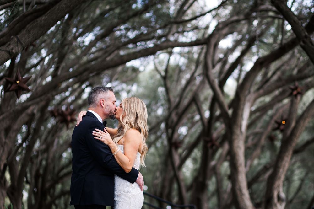 A couple shares a romantic kiss under a canopy of twisted oak trees at dusk.