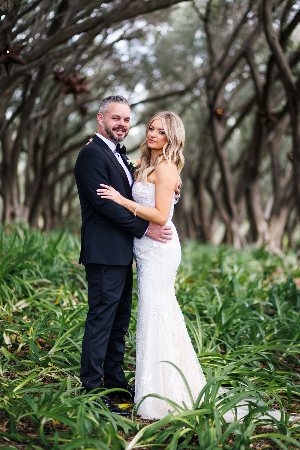 A couple in wedding attire embrace in a lush outdoor setting with arching trees and tall grass.