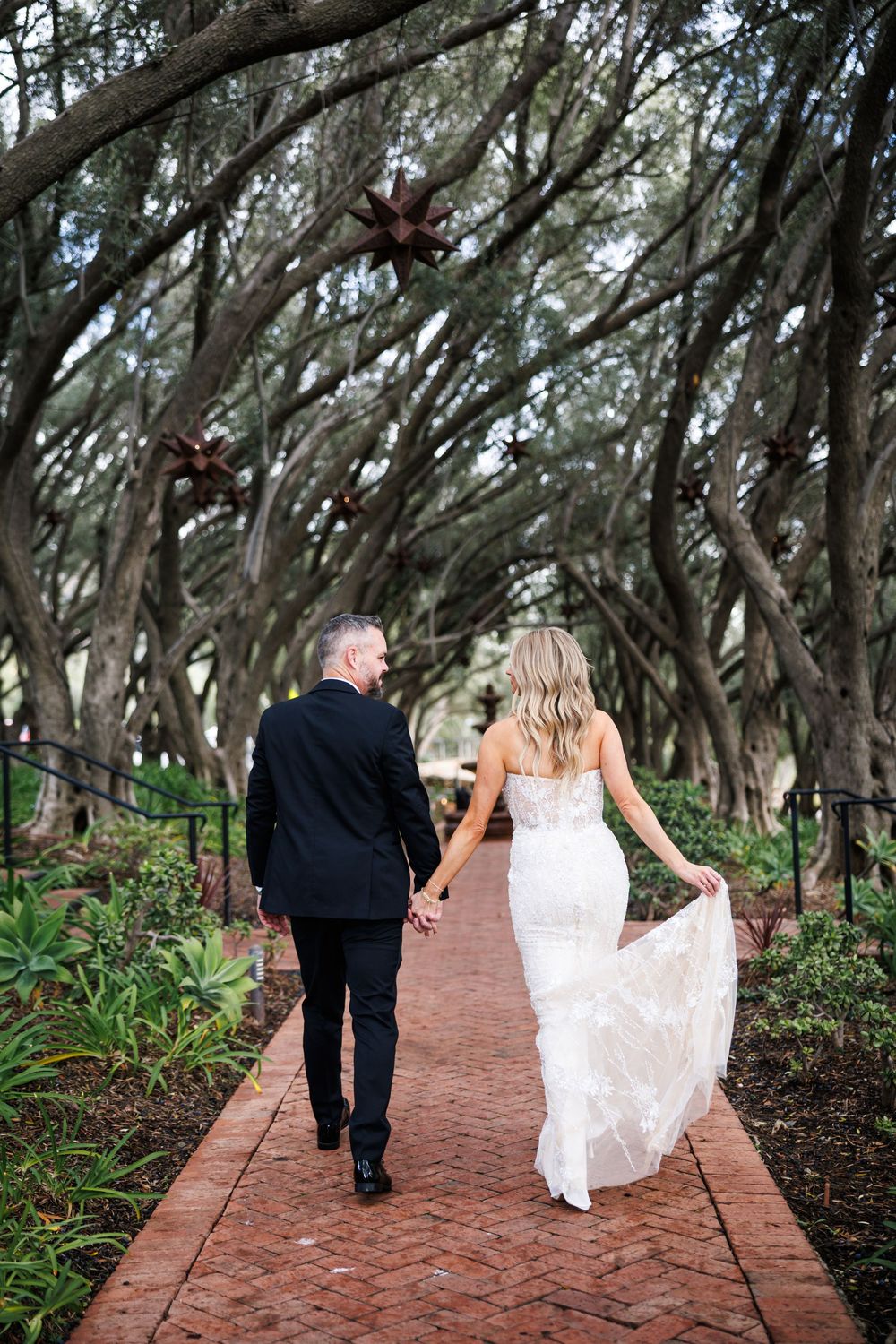 A couple walks along a scenic brick pathway lined with trees and tropical plants.