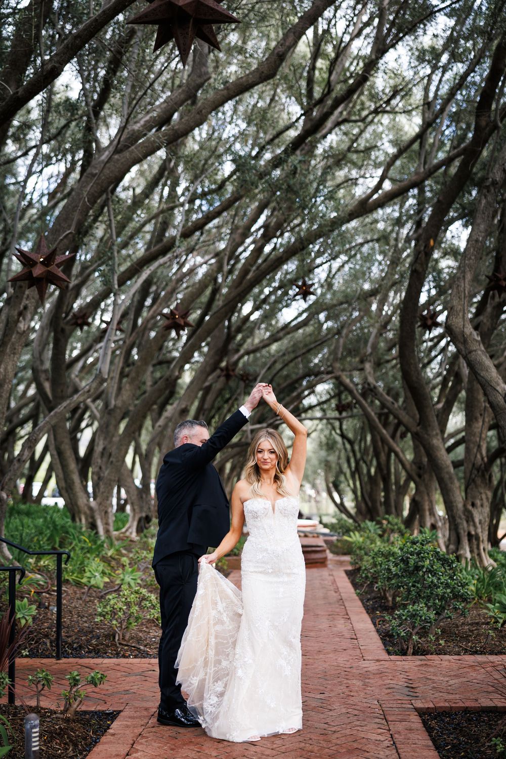 A couple dances and twirls along a brick pathway surrounded by overhanging trees.