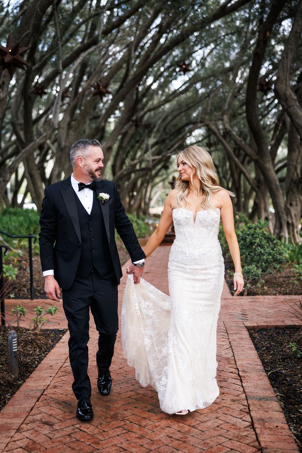 A couple walks along a brick path under a canopy of twisted oak trees in their wedding attire.