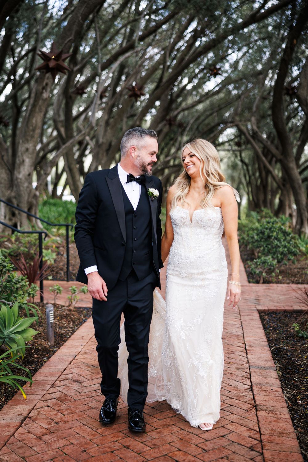 A couple in wedding attire walks along a brick pathway under a canopy of oak trees at an outdoor venue.