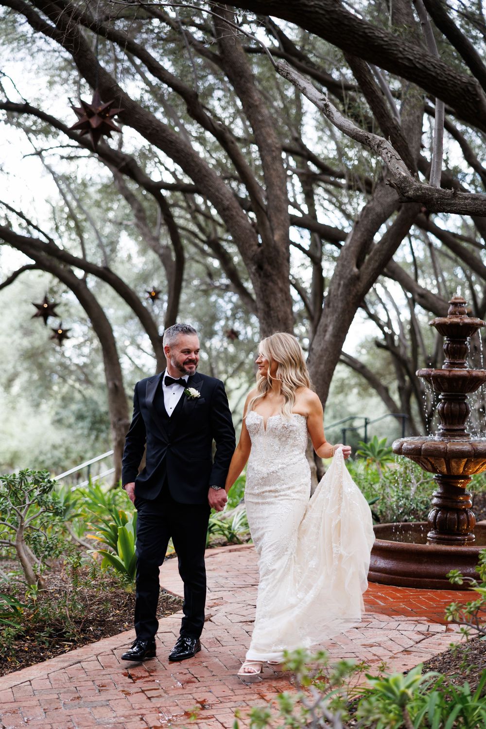 A couple walks along a brick pathway surrounded by trees and a decorative fountain in an outdoor garden setting.