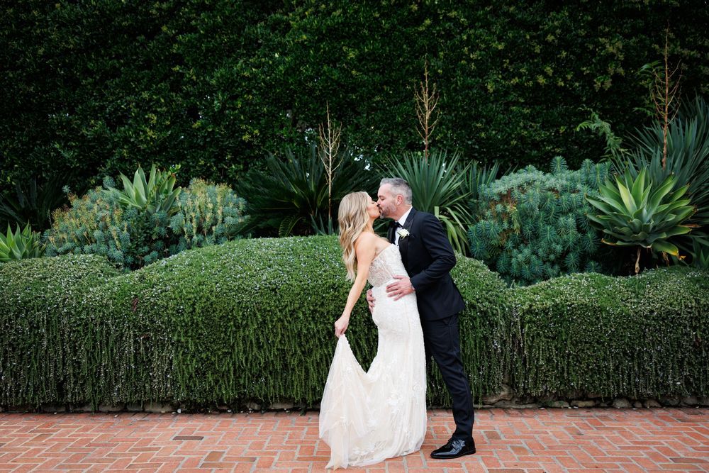 Wedding couple poses in front of lush garden landscaping at sunset.