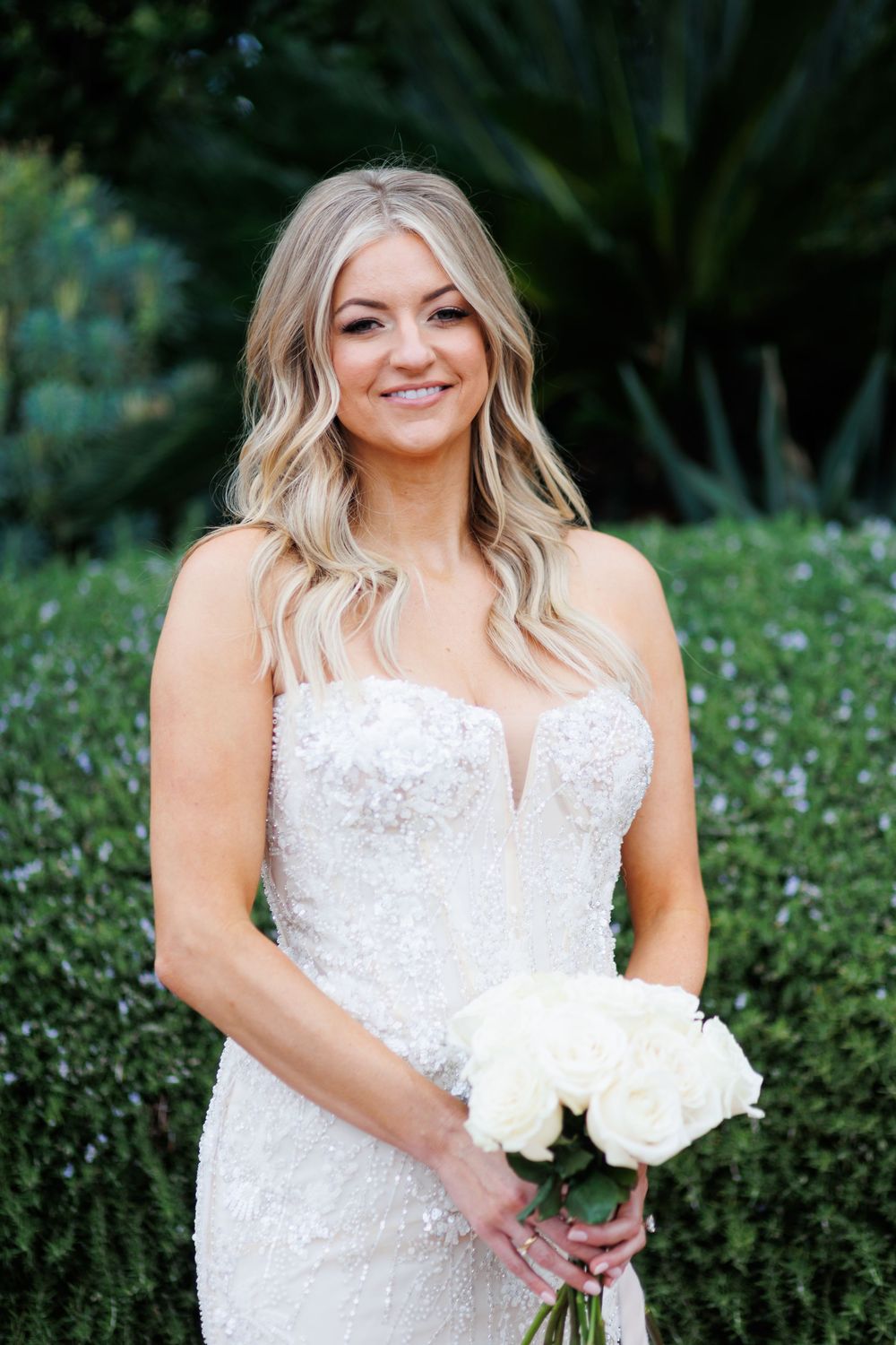 A bride in a white lace wedding dress poses outdoors in a garden setting with white flowers.