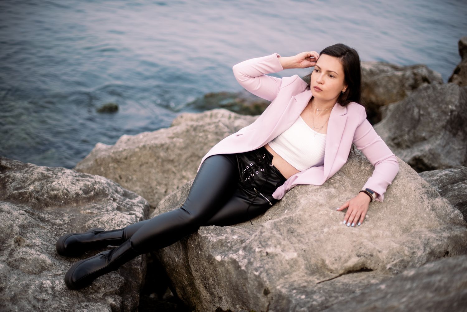 Figure in pink blazer poses gracefully on coastal rocks by the water.