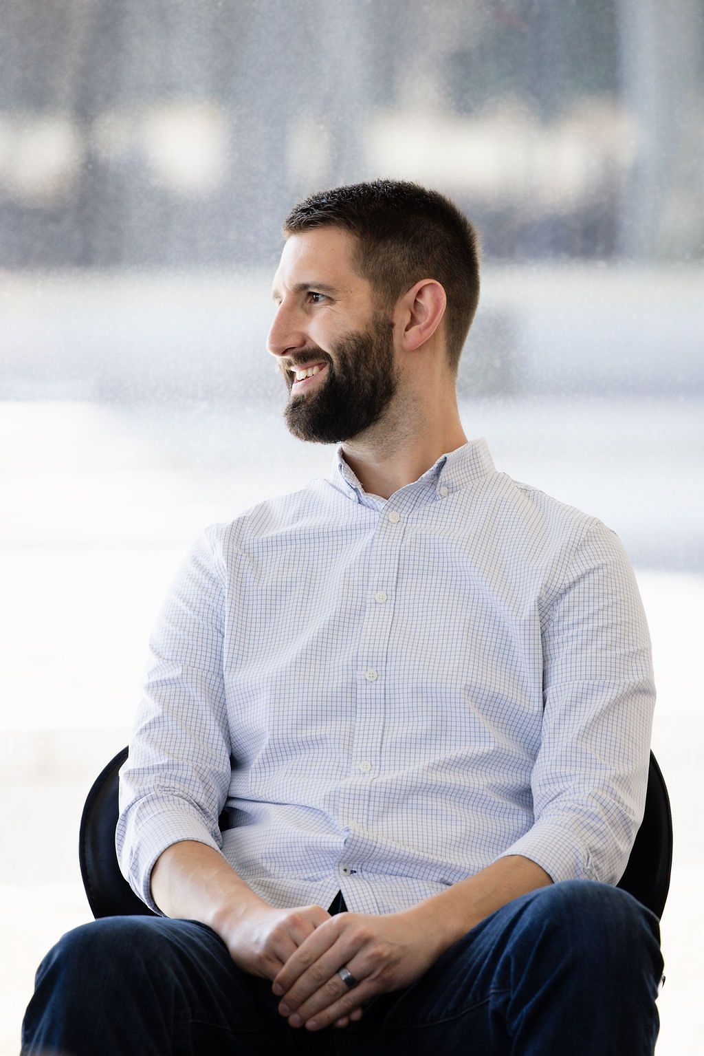A person in a white button-down shirt and jeans sits relaxed in a chair against a bright background.
