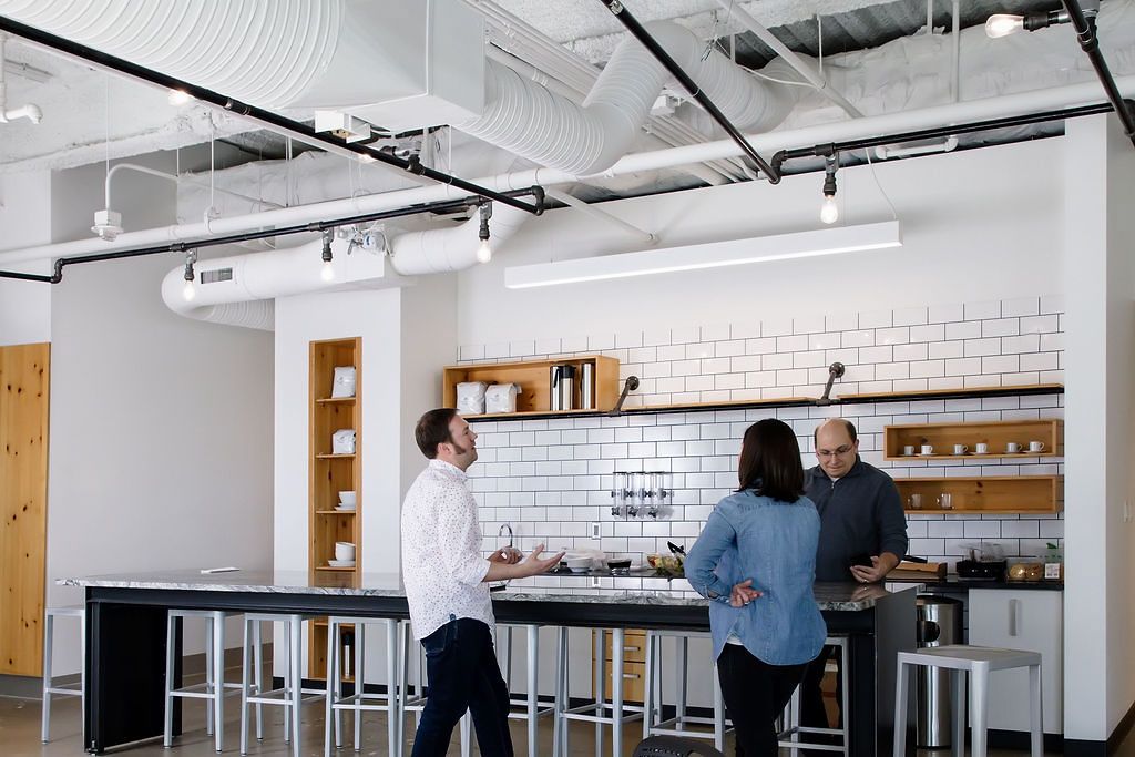 Modern office break room with white subway tile wall, wooden shelving, and people gathering at a counter area.