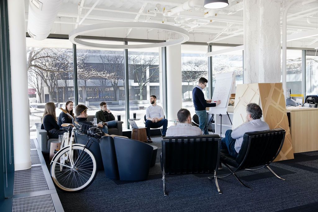 Modern office space with people working at desks, featuring large windows and a bicycle in the corner.