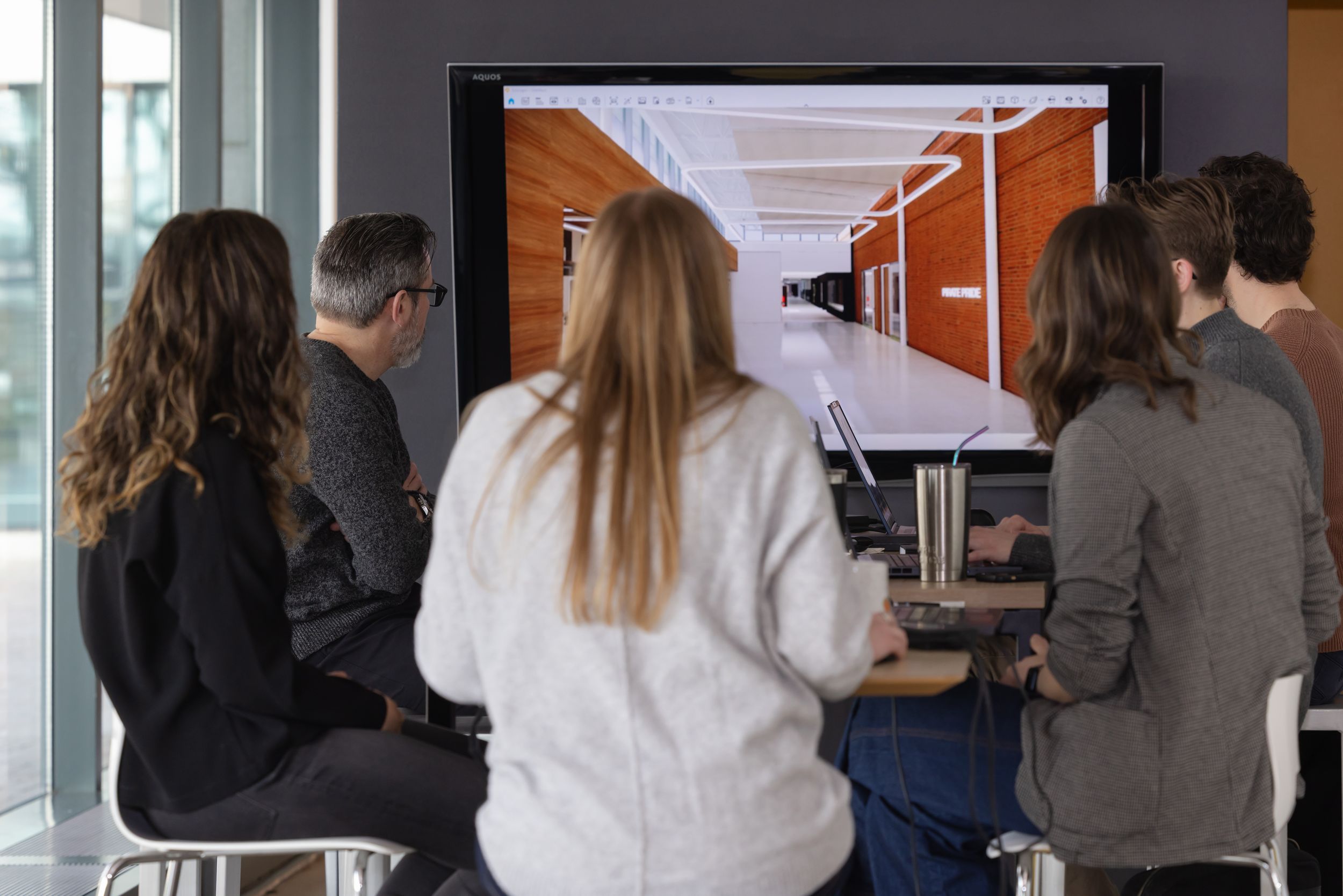 Students gather around displays and monitors in a modern educational space during a presentation or exhibition.