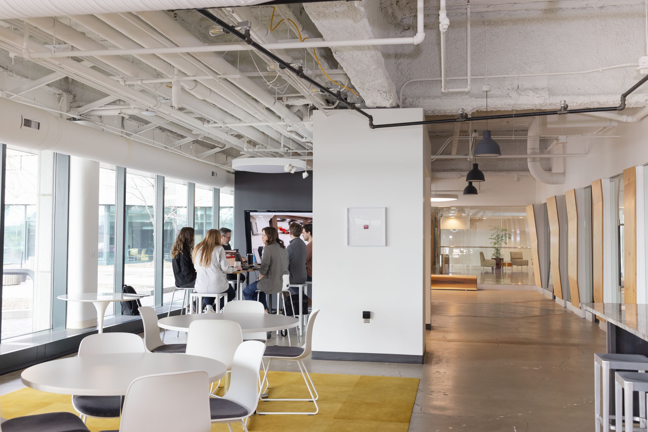 Modern open office space with white walls, exposed ceiling, and large windows showcasing natural lighting.