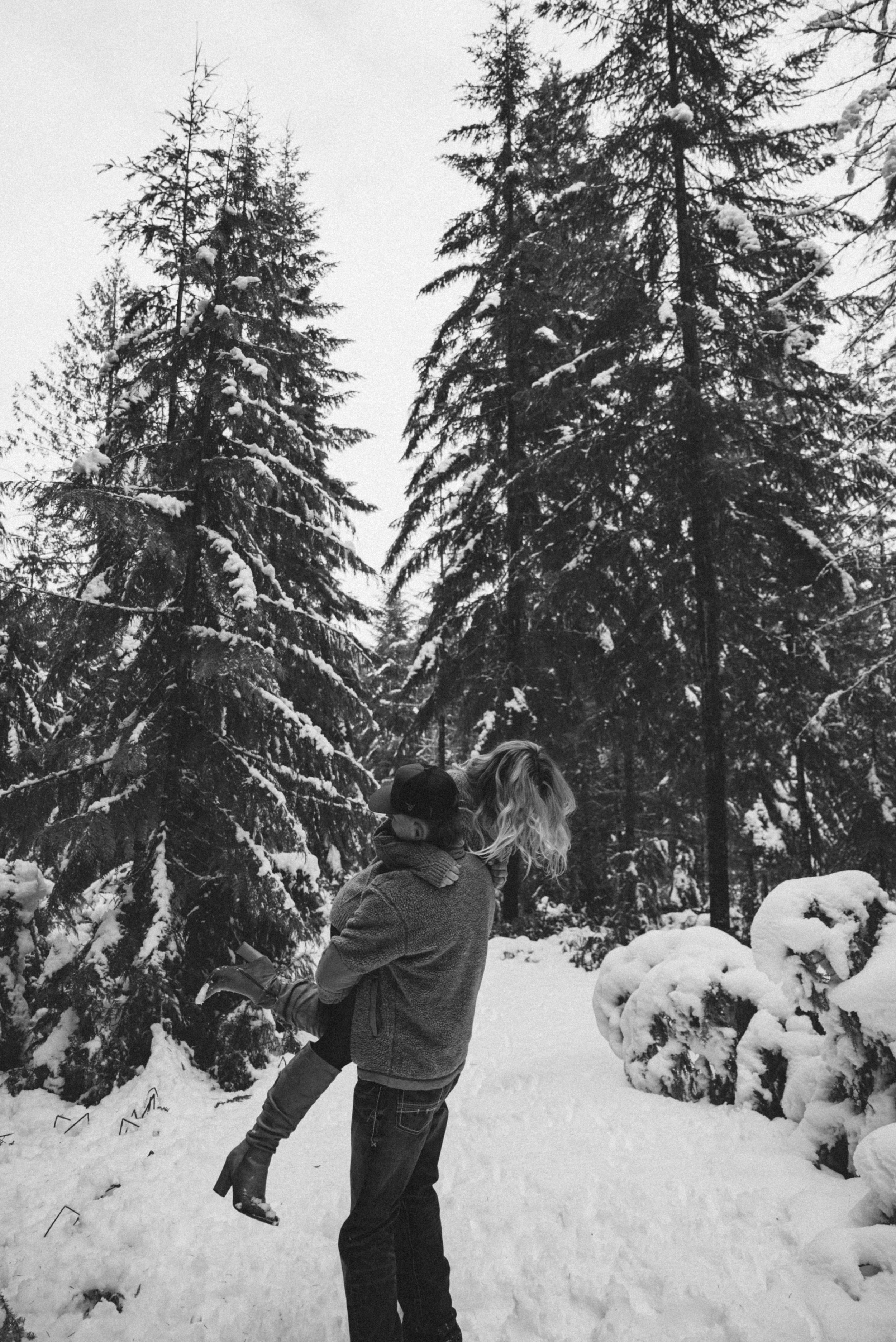 A person throws a snowball in a snowy forest with tall evergreen trees covered in white powder.