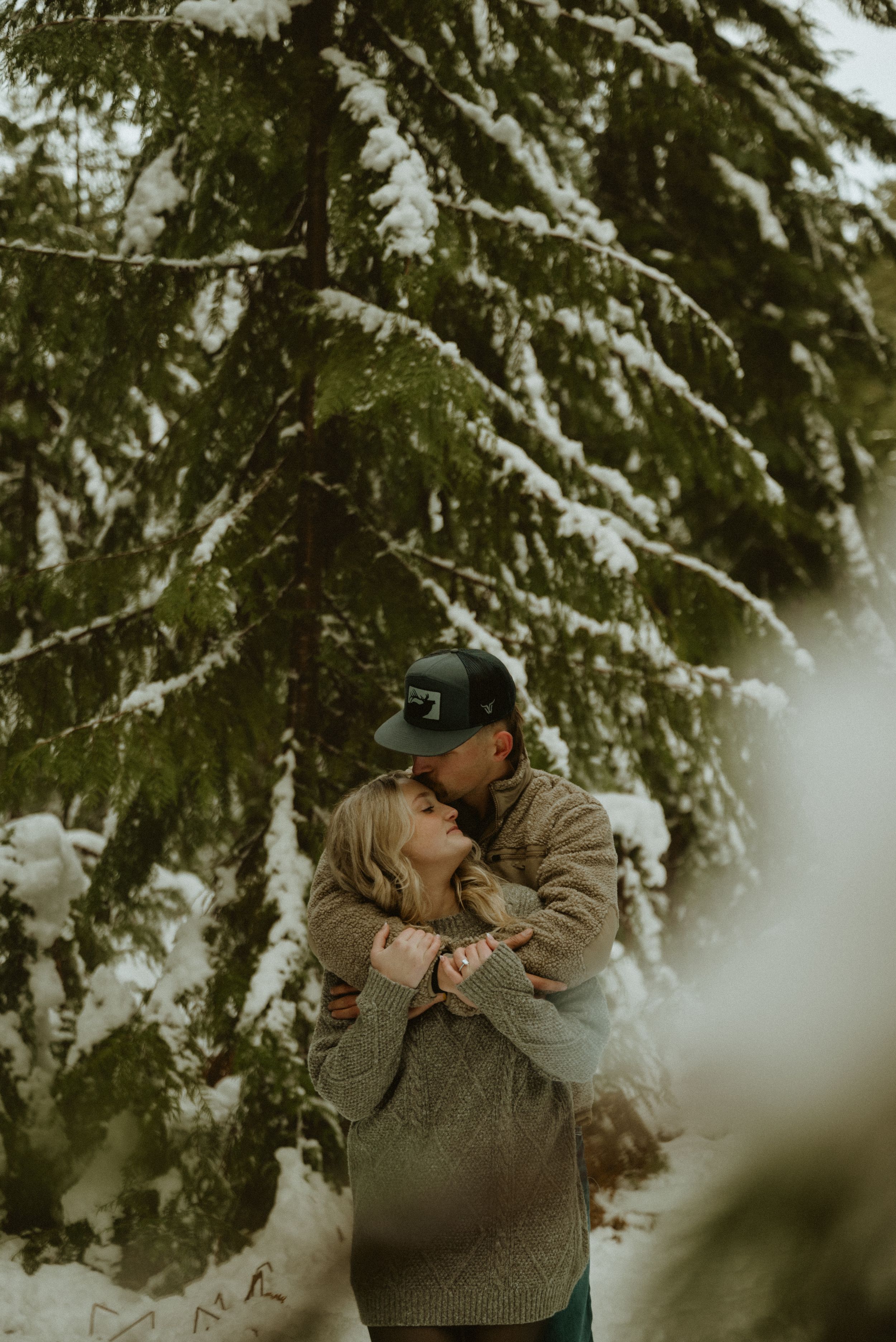 A romantic winter engagement photoshoot in a snow-covered forest with evergreen trees in the background.