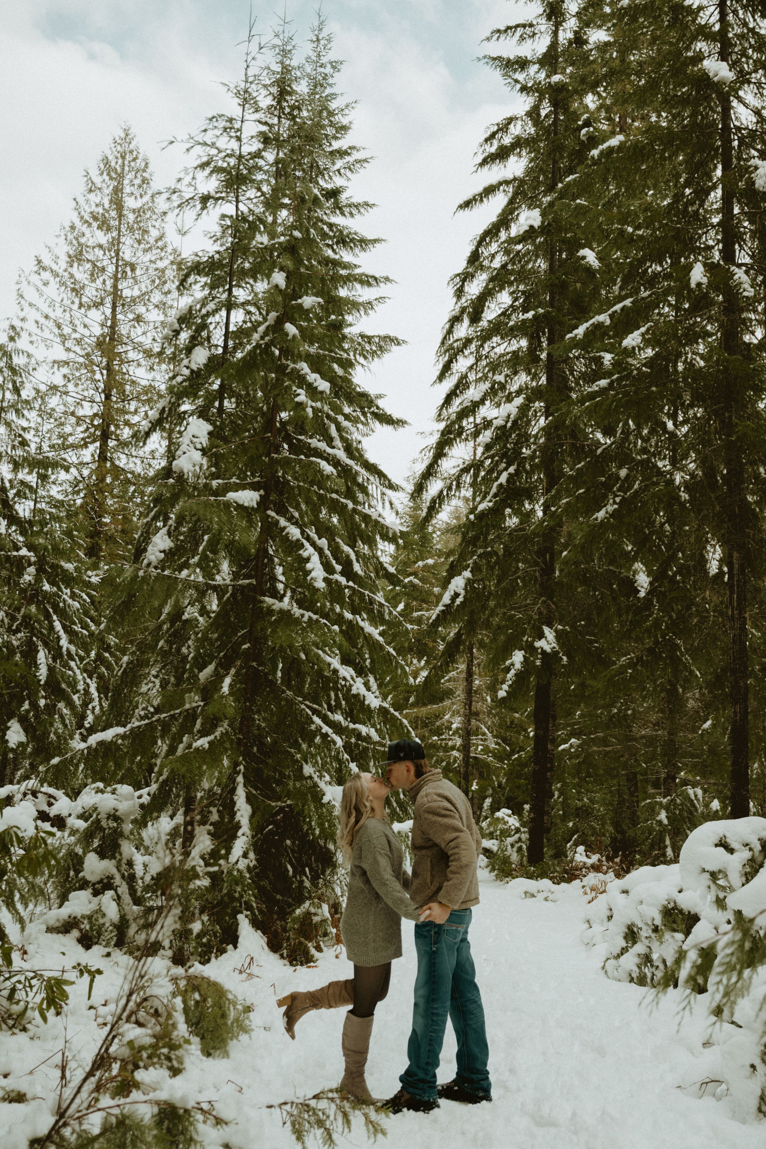 A couple shares an intimate winter moment in a snow-covered forest with tall evergreen trees.