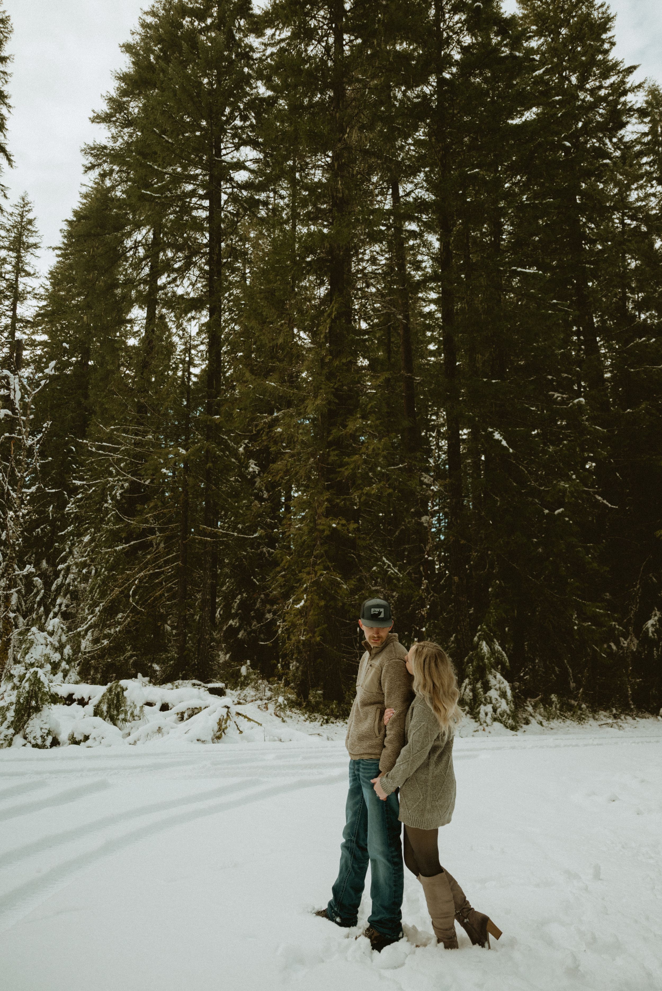 A couple enjoys a romantic winter moment in the snow-covered forest with tall evergreen trees in the background.