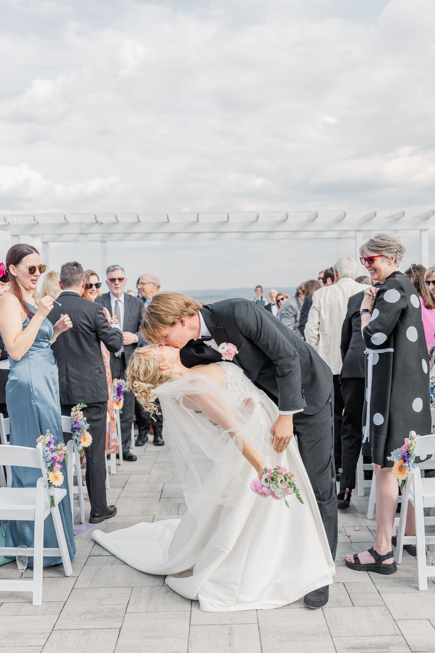 Wedding guests gather on an outdoor terrace during a celebration with white chairs and scenic views.