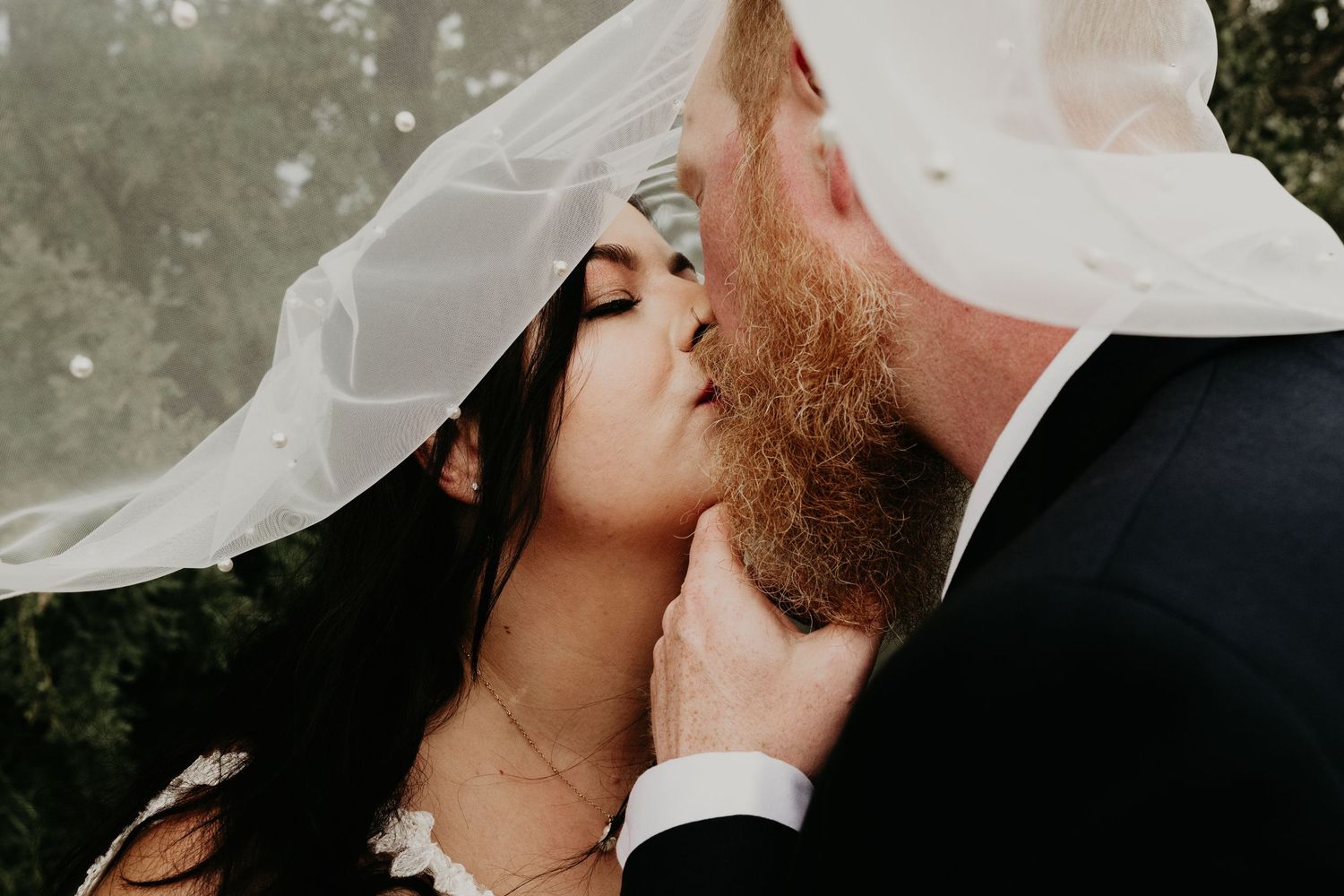 A romantic wedding kiss under a flowing white veil during an outdoor ceremony.