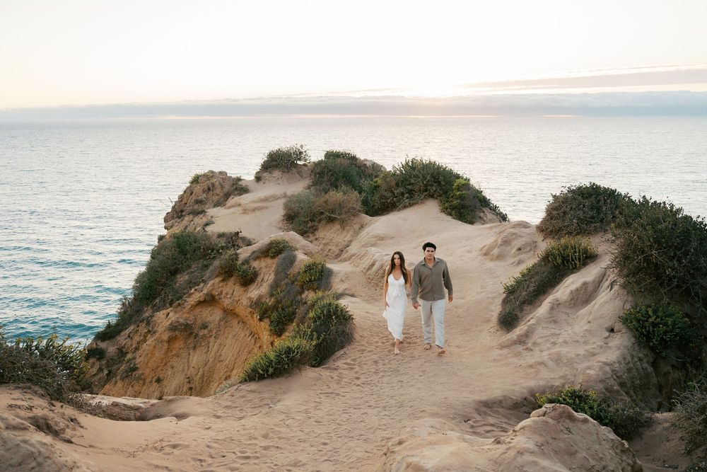 A couple walks along a scenic coastal cliff path overlooking the ocean at sunset.